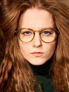 Young Woman With Curly Red Hair in Glasses Looks Directly at Camera