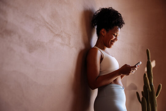 Smiling Woman in Activewear Checking Phone Against Wall