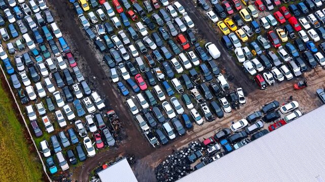 Aerial view of car scrapyard with tire recycling piles. High angle shot of sorted scrapped vehicles next to a warehouse roof and large stacks of old black tires.