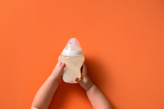 Baby's tiny hands holding a bottle of milk against a vibrant orange