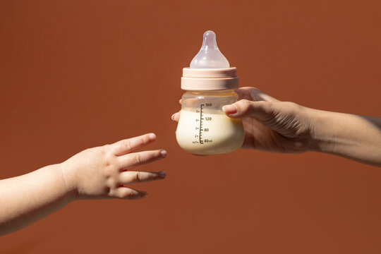 A parent's hand offers a baby bottle filled with milk to a baby's hand