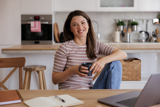 Woman Enjoys Coffee in Kitchen While Sitting on Table working laptop 