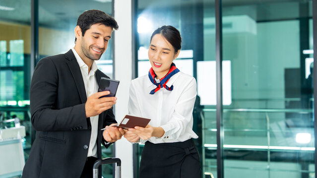 Professional Asian ground staff checking passport for a smiling Caucasian businessman holding smartphone and luggage in a modern airport terminal lobby