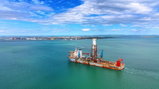 Industrial offshore drilling vessel anchored near coastal city harbor. Aerial close up shot of a specialized drillship with a tower crane on turquoise sea.