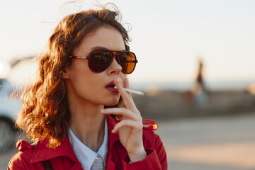 Cheerful woman outdoors, sunglasses on, red jacket, smoking cigarette, sunlit moment, relaxed posture, urban waterfront background, warm tones © SHOTPRIME STUDIO