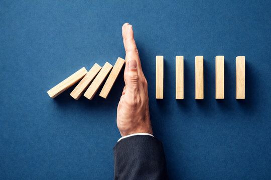 Businessman hand stops falling wooden dominoes on a dark blue surface to demonstrate the prevention of a major risk and the importance of professional intervention for maintaining operational.