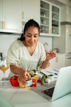 Woman preparing vegetables while following an online recipe on a laptop in a home kitchen