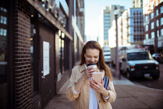 Smiling young woman with coffee using smartphone on city sidewalk