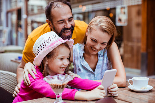 Happy parents and daughter using smartphone at outdoor cafe