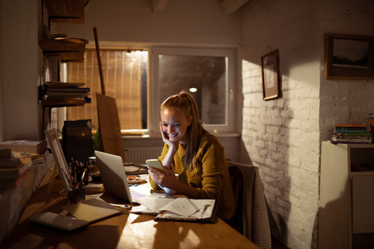 Smiling young woman using smartphone while working late at cozy home office