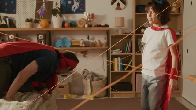 Father and young son wearing superhero capes navigating yarn laser maze in bedroom. Ideal for promoting family bonding, creative parenting, and active indoor play
