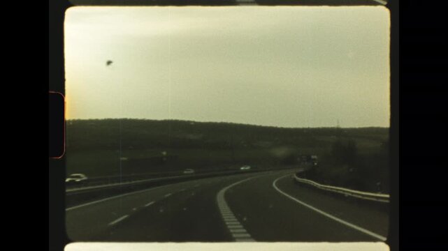 Point of view of driving along an elevated highway in the rain passing an overhead directional road sign under a grey overcast sky captured on retro super 8 film