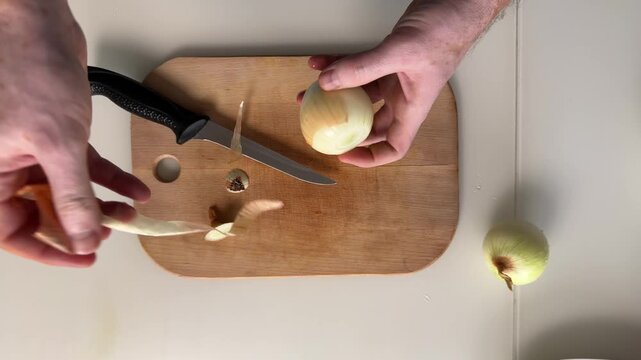 Top-down view of hands preparing halved onion on wooden cutting board