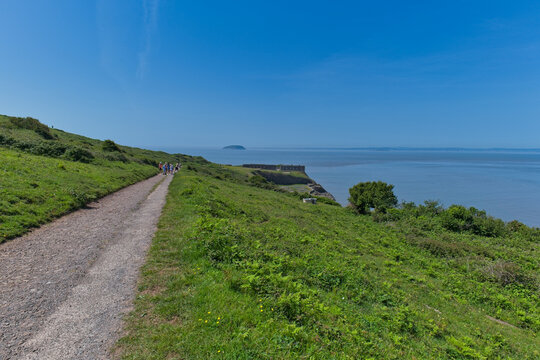 A scenic coastal path winds along green, grassy cliffs under a clear blue sky, overlooking the sea with a distant island on top of Brean Down - Burnham-on-Sea - UK