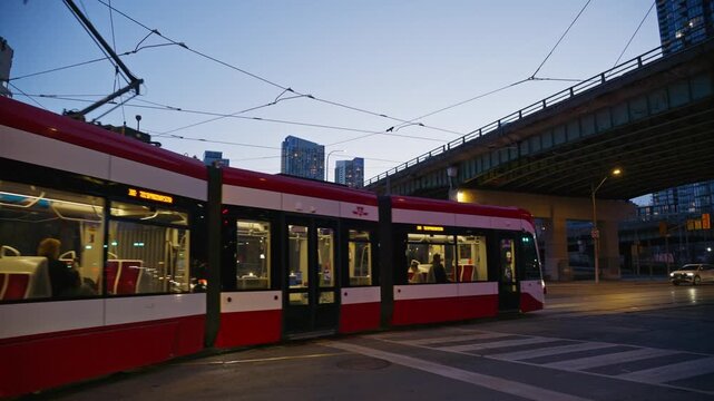 Toronto streetcar moving through urban intersection during twilight with city buildings, downtown traffic, commuters on public transport and illuminated Gardiner Expressway