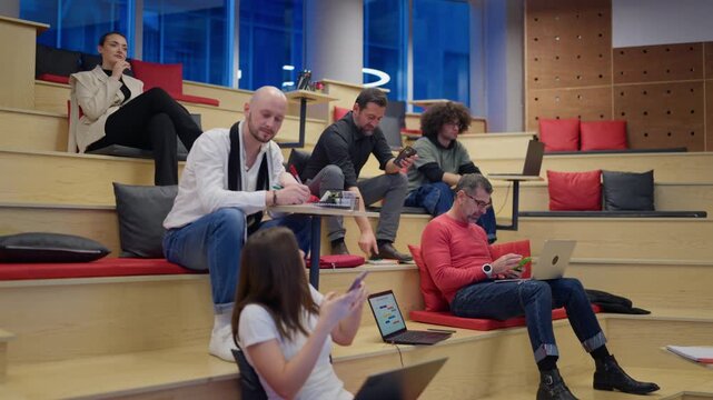 Diverse professionals are working and interacting in a modern open-plan office. Coworkers utilize technology and notebooks in a tiered workspace