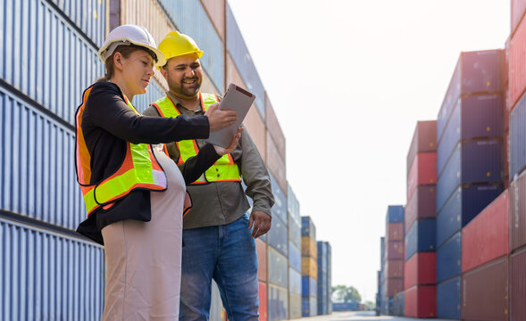 Female pregnant manager and male engineer reviewing tablet digital data shipping container cargo yard port logistics  with safety helmet and high visibility vest