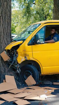 A severely damaged yellow commercial van rests near a large tree after a collision. A man examines the wrecked front end and scattered boxes in an outdoor setting.