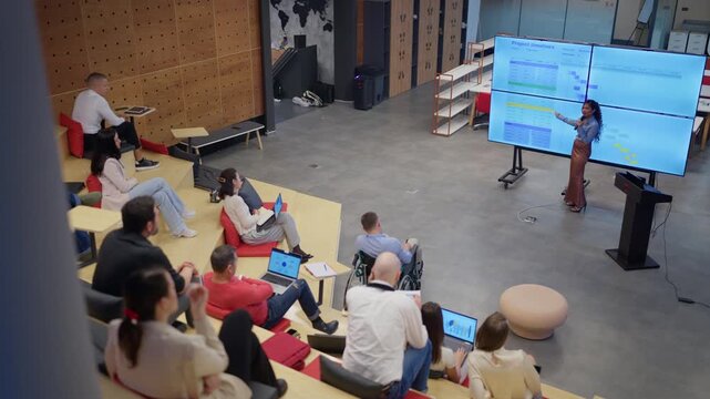A businesswoman delivers a presentation on project timelines to a diverse group of professionals. Her colleagues are attentively listening during a corporate meeting in a modern office