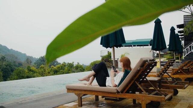 Woman relaxing on lounge chair using smartphone by infinity pool
