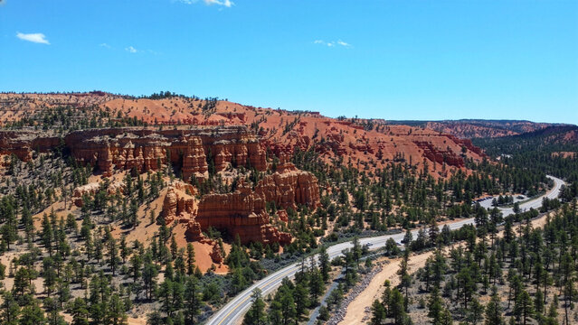 Breathtaking bird's-eye view of a modern asphalt highway cutting through lush green valleys and forests in Dixie National Forest, Utah. Captured on a bright sunny day under a clear blue sky.