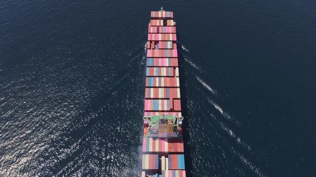 High-angle aerial perspective following a massive container ship as it sails across the deep blue sea, showcasing the scale of global maritime trade and freight transportation logistics