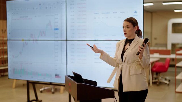 A female professional presents financial data and charts on large screens during a business meeting. An expert employee is delivering a clear presentation in a modern office environment