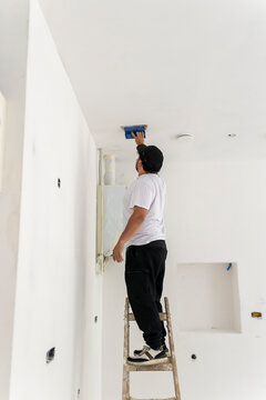 Construction professional standing on ladder while smoothing ceiling plaster in unfinished apartment, safe renovation workflow and drywall finishing