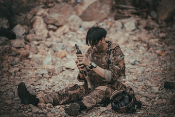 Obraz premium Exhausted tactical military soldier sitting on rocky terrain, deeply focused on inspecting his rifle weapon after an intense combat mission. Fatigue and warfare survival concept.