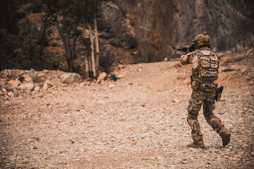 Obraz premium Armed military operator advancing through rugged rocky terrain while aiming a rifle during a combat patrol mission. Tactical soldier moving forward in dangerous combat zone concept.
