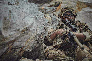 Obraz premium Armed tactical military operator taking cover behind large rocks during combat mission. Soldier with rifle holding defensive position in rugged terrain. Warfare and survival concept.