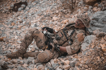 Obraz premium Exhausted military commando resting on rocky terrain during a combat mission. Tired armed forces operator catching his breath after battle. Tactical survival and warfare concept.