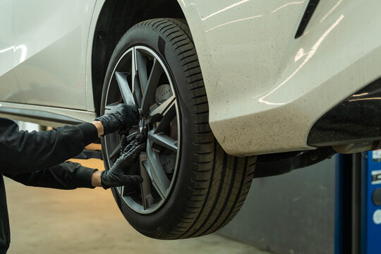 Gloved hands work on a car wheel while the vehicle is raised on a lift inside an auto repair workshop, with close up detail of the rim, tire, and underbody in