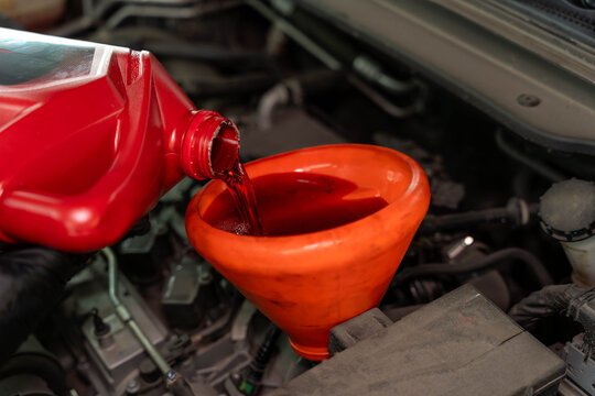 A close up view of motor oil being poured from a red container into an orange funnel inside a car engine bay, with hoses, engine parts, and workshop lighting