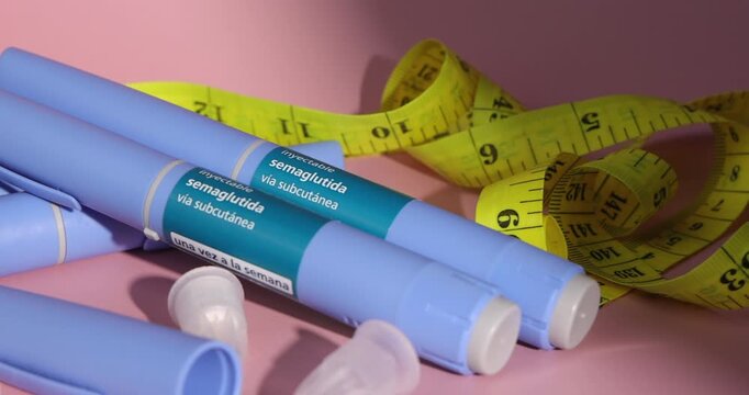 Horizontal pan over various semaglutide glp1 medication pens with extra replacement needles and a yellow tape measure on pink gradient.