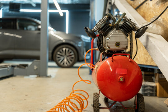Red portable air compressor with coiled orange hose in a garage workspace, with a blurred car in the background, industrial lighting, and tools suggesting