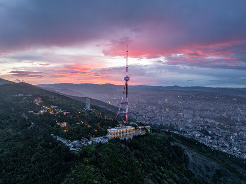 Vibrant sunset over Tbilisi TV tower and Mtatsminda park from above, high angle drone view of illuminated city landmark building and ferris wheel at dusk with pink clouds, Georgia.