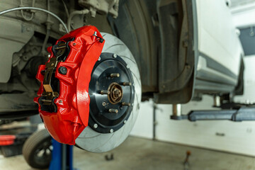 Close view of a bright red brake caliper mounted on a ventilated brake disc and wheel hub in an auto repair workshop with a vehicle lifted in the background © Egor Lint