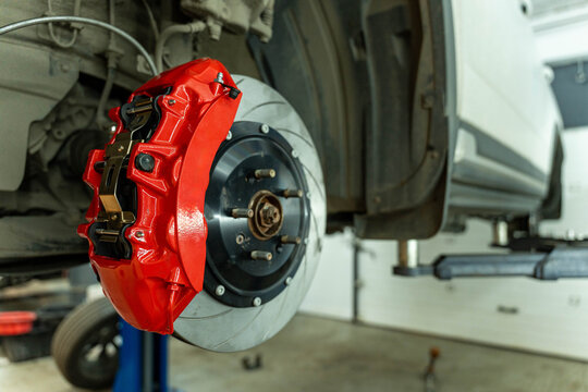 Close view of a bright red brake caliper mounted on a ventilated brake disc and wheel hub in an auto repair workshop with a vehicle lifted in the background