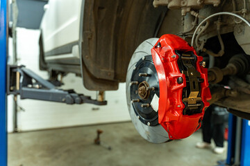 Close view of a car wheel hub with a bright red brake caliper and rotor exposed during maintenance in an auto repair workshop, with the vehicle lifted and a © Egor Lint