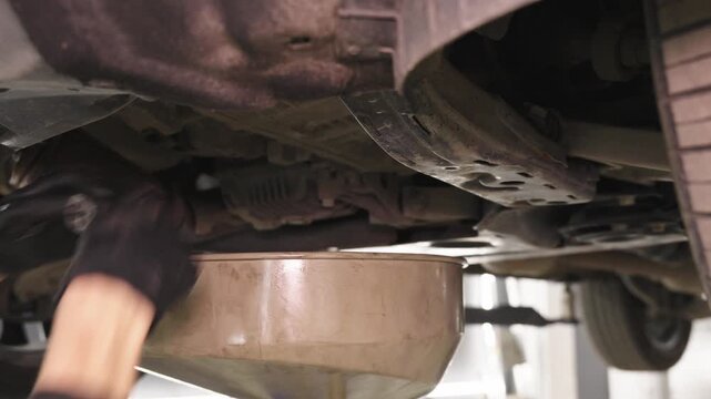 A mechanic reaches beneath the front of a lifted car during maintenance, with a drainage container placed below the undercarriage in a bright workshop setting