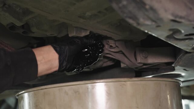 A black-gloved hand works under a lifted vehicle as used oil drains into a metal pan in a dim garage, capturing routine automotive maintenance from a low angle
