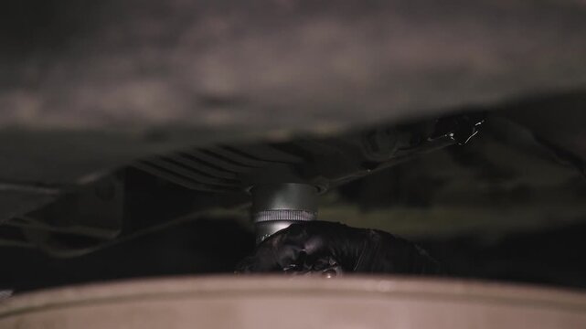 Tight low-angle view beneath a car reveals dark mechanical components, metal parts, and structural elements with shallow focus in a dim workshop setting