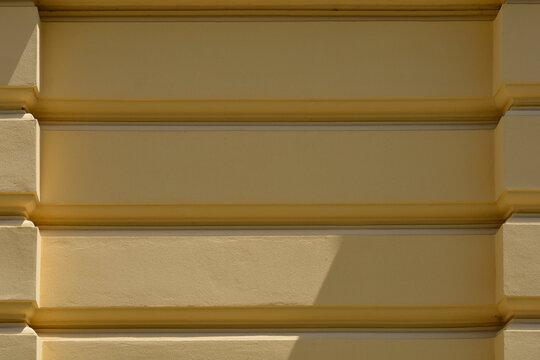 An abstract architectural detail of a yellow painted wall with strong horizontal rusticated plaster lines and shadow play