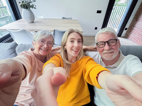 Young woman taking a joyful selfie with her smiling grandparents on a cozy living room couch, pointing at the camera and enjoying relaxed multigenerational family time together in a bright modern home