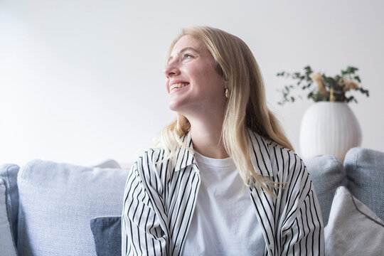 Young woman sitting on a light sofa smiling and looking up, relaxed at home in casual striped shirt and white tee, cozy living room with soft pillows and decorative vase peaceful, hopeful mood.