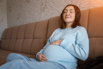 Obraz premium A pregnant woman sits comfortably on a home sofa with her hand resting on her belly. She wears a light blue sweater and relaxes with her eyes closed. Warm natural sunlight fills the cozy living room