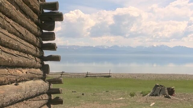 Gaze upon majestic Lake Khuvsgul in Mongolia, framed by a rustic log cabin. Serene waters reflect a cloudy sky, with distant mountains gracing the horizon.