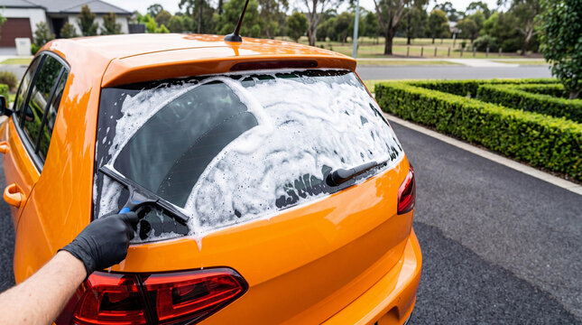 Car detailer using squeegee to clean foam from rear window of orange vehicle. Professional mobile car wash service process. Automotive maintenance and vehicle care industry concept.