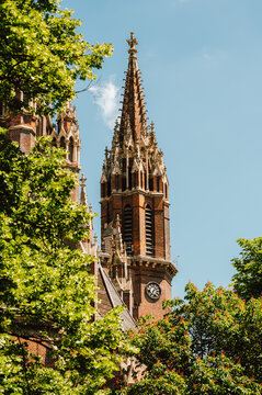 Tall Gothic church tower with a richly detailed brick spire and decorative pinnacles rising above surrounding leafy trees on a clear blue sky day. Visible